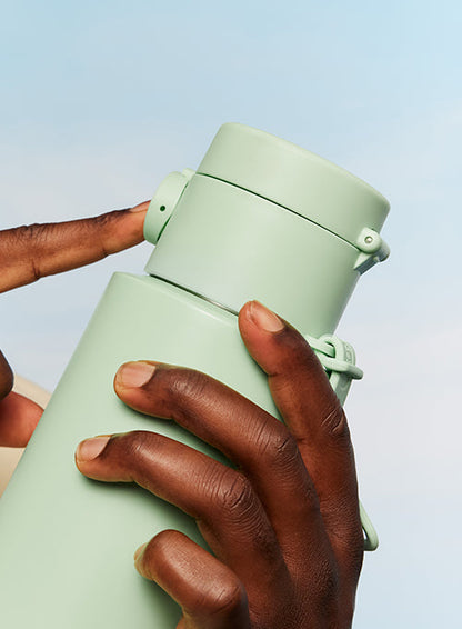 A close-up of a person's hands holding a light sage green vacuum-insulated water bottle against a pale blue sky background.
