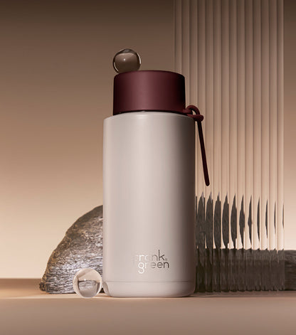 A frosted light gray insulated bottle with a dark burgundy lid and matching carry loop, standing on a beige surface with a stone and ribbed panel in the background. The bottle shows the Frank Green logo near the bottom.