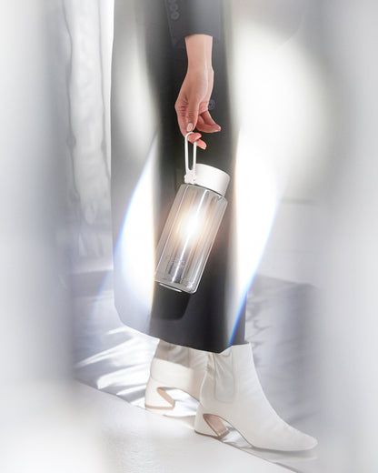 A person in a black coat and white ankle boots holds a clear cylindrical water bottle with a white cap and loop handle, lit by a warm glow from within.