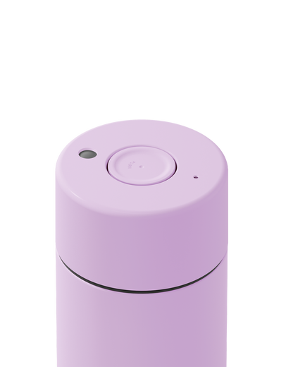 Close-up of the top of a matte lavender cylindrical container, showing a circular lid with a central button and small side openings, on a white background.